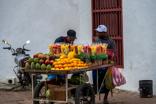 Cartagena, Columbia — Jan 11, 2023. A pair of merchants stands by their food cart carrying a colorful display of fruits in a court yard.