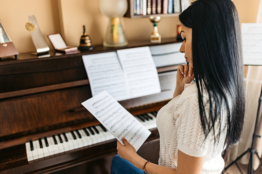 Beautiful young woman looking at her musical sheet before playing her piano in her room at home.