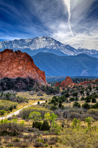 Image of the Garden of the Gods park in Colorado.  In the foreground there are green trees and bushes and Pikes Peak can be seen in the background against a stormy sky.