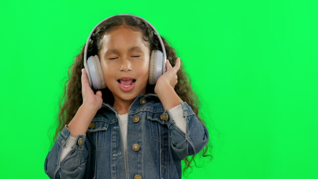 Little girl, dancing and singing with headphones on green screen against a studio background. Happy female person, child or kid listening to music and enjoying dance, audio or sound track on mockup