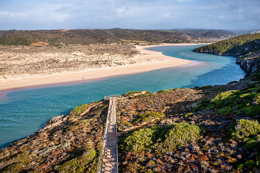 Aerial view of Wooden walkway to the beach Praia da Amoreira at river Ribeira in Aljezur, snaking through the golden sand of Praia da Amoreira beach with crystal clear calm water, sand dunes and clouds in the sky on a sunny day in October.