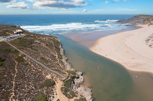 Aerial view of Wooden walkway to the beach Praia da Amoreira at river Ribeira in Aljezur, snaking through the golden sand of Praia da Amoreira beach with crystal clear calm water, sand dunes and clouds in the sky on a sunny day in October.