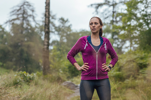 Three quarter length of a mature woman wearing sports clothing going for a run in the woods to keep fit on a rainy day at Hollows Farm, The Lake District in Cumbria, England. She is standing with her hands on her hips, looking away from the camera.