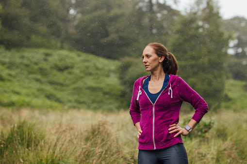 Three quarter length of a mature woman wearing sports clothing going for a run in the woods to keep fit on a rainy day at Hollows Farm, The Lake District in Cumbria, England. She is standing with her hands on her hips, looking away from the camera.
