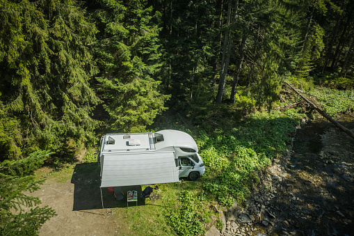 Aerial View of White Camper Van Parked Solo in the Forest Next to Woodland Stream. RV Camping and Traveling Theme.