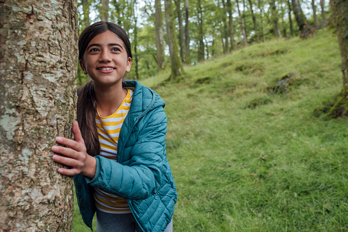 Three quarter length of a girl walking down the side of a hill in the forest, she is standing behind a tree on the grass looking away from the camera while on a trip at Hollows Farm, The Lake District in Cumbria, England. She is wearing a coat.