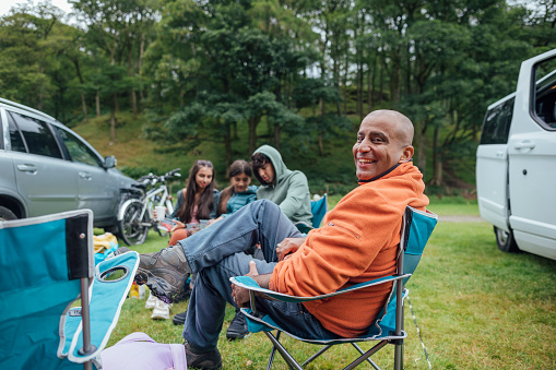 Full length of a small group of siblings sitting together on camping chairs smiling and talking, they are camping and have set up some tents to create a campsite at Hollows Farm, The Lake District in Cumbria, England. They are sitting with their father, he is looking over his shoulder at the camera with a smile.