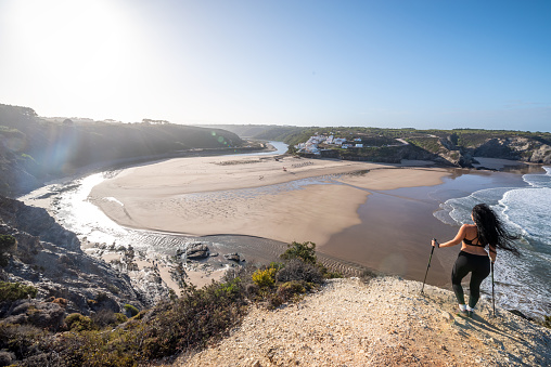 Traveler woman stands on the edge of a cliff near the town of Odeceixe, Algarve region, Portugal watching the sun rising on the horizon.