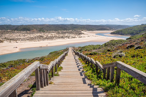 Wooden walkway to the beach Praia da Amoreira at river Ribeira in Aljezur, snaking through the golden sand of Praia da Amoreira beach with crystal clear calm water, sand dunes and clouds in the sky on a sunny day in Octobe