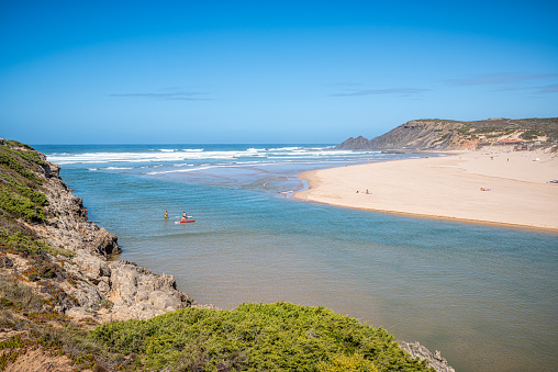 Unrecognizable people swiming and paddle surfing at Praia da Amoreira, one of the surfer's favorite beach with beautiful wide sandy beach, long waves, river and cliffs, Algarve, Portugal