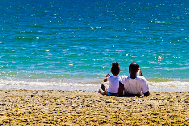 Black family and azure sea father and daughter sit on the beach and look at azure sea girls laying on beach stock pictures, royalty-free photos & images