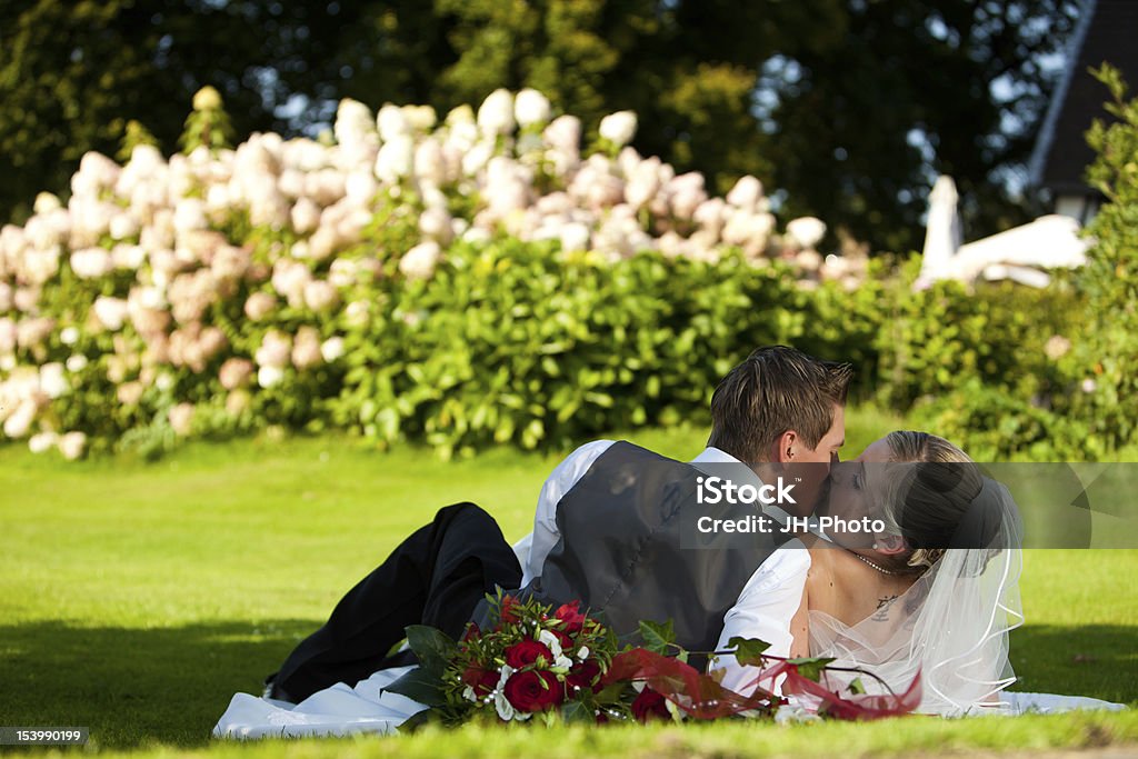 wedding kissing couple on meadow Just married young bride and groom are laying down on the green grass with nice colorful flowers in the background and the bouquet in the foreground. Adam Groom Stock Photo