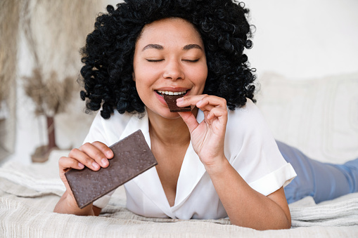 happy african american woman eating milk chocolate with closed eyes while lying in bed and feeling pleasure. smiling woman bite cocoa candy bar and enjoy taste