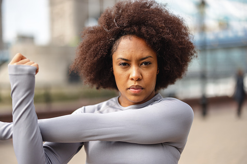 Close-up side shot with blurred background of an African-American mid-adult woman with afro hair warming up while looking serious at camera before exercising as a part of her daily routine before going to the office.