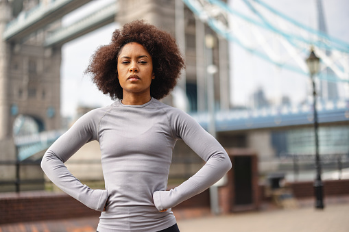 Waist-up side shot with blurred background of an African-American mid-adult woman with afro hair warming up while looking serious at camera before exercising as a part of her daily routine before going to the office.
