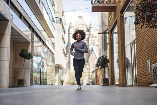 Full length front shot with blurred background of an African-American woman runner practicing outdoors as a part of her daily morning routine before going to the office.