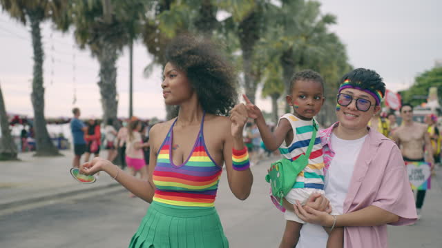LGBTQ lesbian parents and little child walking on road near beach, Multiracial family having cheerful moment in pride parade event, LGBTQ family spending wonderful time outdoors together