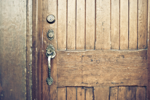 A selective focus image of a worn wooden door with old and new dead bolt locks and an old iron latch handle