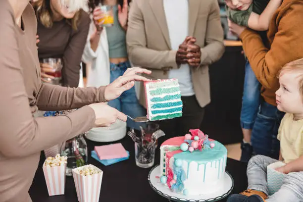 A young Caucasian expectant mother holding a slice of blue cake in the air to reveal the baby's gender. She and her husband are having a boy. The expectant mother is surrounded by her supportive friends and family. They all look happy and excited.