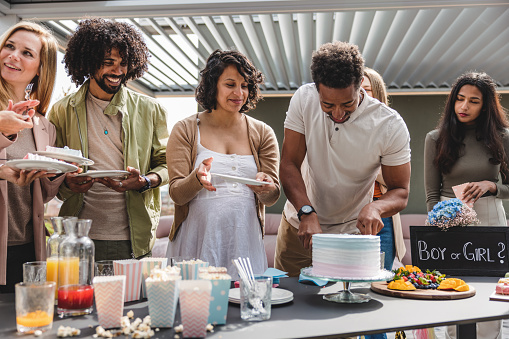 A Black adult father-to-be cutting the cake at a gender reveal and distributing it among the guests. They all look happy and are patiently waiting for their piece of cake. The expectant couple is about to have a baby girl since the cake is pink inside. Beautifully organized outdoors gender reveal party.