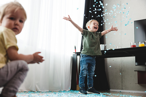 A young caucasian boy playing with blue confetti at a gender reveal party while his younger brother is looking at the camera. Siblings having fun and throwing around confetti.