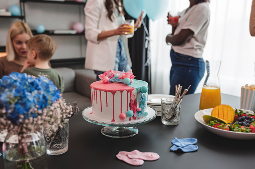 Beautiful delicious pink and blue cake on a cake stand at a gender reveal party. The table is full of other delicious snacks such as fresh fruits and popcorn. In the background, there are some party guests that are catching up and having fun conversations.