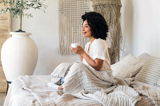 african american woman covered with blanket drink hot tea feeling pleasant and smile. morning weekend at home. concept of recreation on holidays. young female enjoy cup of coffee in bed