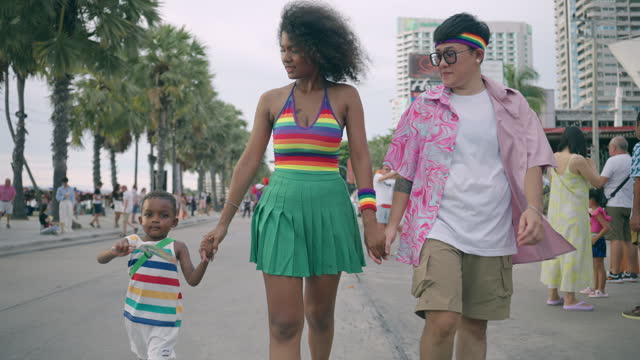 LGBTQ African mom and LGBTQ Asian dad walking and holding African son hands in pride parade event, LGBTQ lesbian parents and little child having wonderful time as happy family, Multiracial family supporting diversity