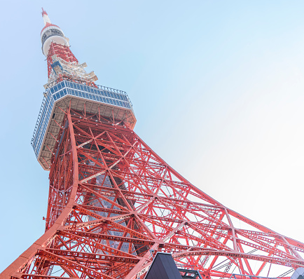 The Tokyo Tower, an iconic landmark, stands tall in the heart of Tokyo, Japan, against a clear blue sky. the iconic Tokyo Tower standing tall against the clear blue sky, dominating the urban landscape with impressive grace. The tower, painted in a striking combination of orange and white, extends upwards from its base, with its pointed antenna reaching even higher. The image offers a captivating view of the tower in its full glory, with the bustling cityscape of Tokyo in the background. The tower's leg extends vertically towards the base, with a series of observation platforms gradually widening as they climb higher. The upper half of the tower is complemented by a distinctive, three-pronged antenna structure, which projects above the observation deck. A stunning close-up of the tower's intricate detail and elaborate lines is also featured, showcasing the tower's fascinating architecture in great detail, from its geometric shapes to its diagonal lines and carefully-placed panels.