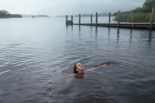 Woman swimming in a lake in Keswick, the Lakes. He has his eyes closed as he enjoys the cold water dip as it rains.