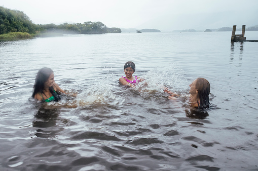 Mother and her two daughters open water swimming together in a lake in Keswick, the Lake District. They are on a staycation enjoying time together, splashing each other.