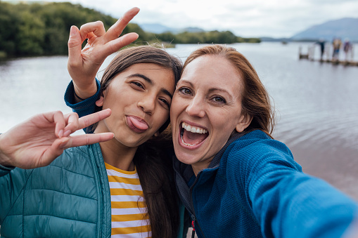 Woman and her young daughter taking a selfie while on a hike outdoors in Keswick, Lake District. They are going open water swimming at a lake. They are smiling looking at the camera, doing peace signs while pulling faces.