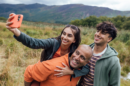 Father, daughter and son on a walk together through the countryside in Keswick, The Lakes. They are using a smart phone to take a selfie while the father gives his daughter a piggyback.