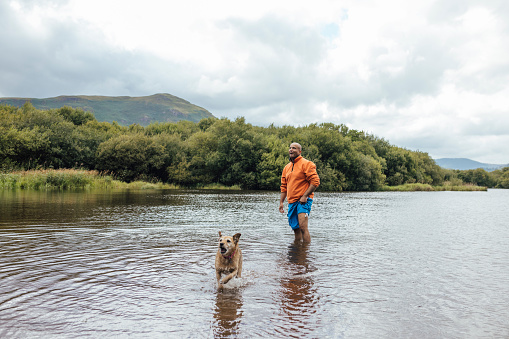 Man standing in a lake while on a hike outdoors in Keswick, the Lake District. He is walking his dog who is excited running through the water.