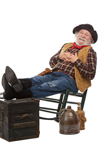 Smiling old cowboy sits in rocking chair with feet up Classic old west style friendly cowboy with felt hat, grey whiskers, revolver. He leans back in a rocking chair with feet up. Isolated on white, vertical, copy space. checkered cowboy boots stock pictures, royalty-free photos & images