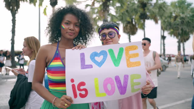 African-Asian LGBTQ lesbian couple showing banner to emphasize diversity and human rights in pride parade event, LGBTQ lesbian couple holding love is love banner, Multiracial couple spending time outdoor together near seaside