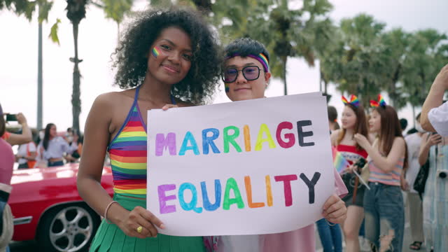 African-Asian LGBTQ lesbian couple holding banner to show marriage equality and human rights during participating in pride parade event, Multiracial couple having cheerful time outdoor together near seaside