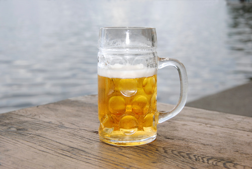 close up of a beer mug with fresh beer with foam and bubbles in a beer garden in front of a lake