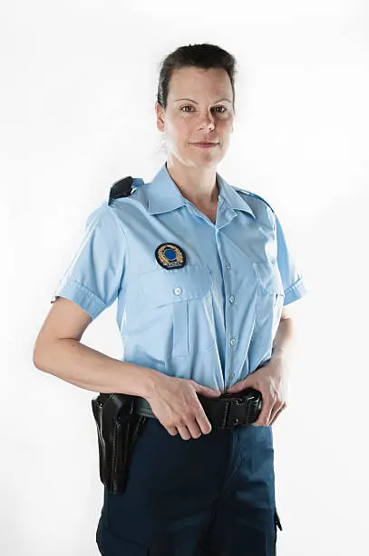 Policewoman standing on white background. She is wearing a summer blue shirt and her hand are resting on her gun belt. She is looking at camera with a light smile. American shot with studio lighting, vertical with copy space. 