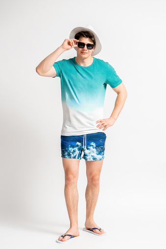 Young dark-haired man in a white hat, beach outfit and flip-flops adjusting his sunglasses posing full length in the studio on a white background