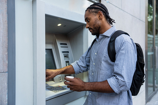 Side view of African American man using ATM while holding plastic cup