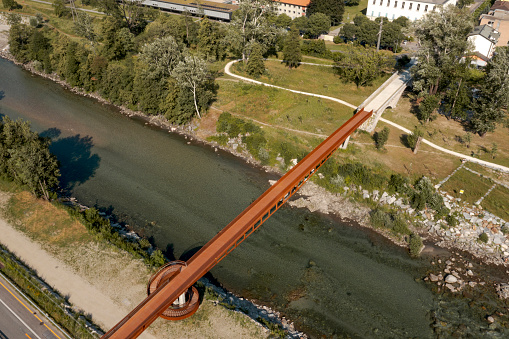 Modern rusted pedestrian and bicycle bridge with flowing Ticino river below. Beautiful panorama of the Alps of Italian Switzerland, taken from the sky. No one inside