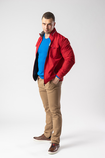 A man of athletic build in a red jacket, blue t-shirt and beige pants stands and holds his hands in his pockets. Model posing on a white background.