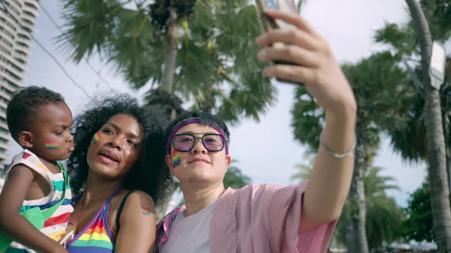 LGBTQ multiracial family taking a selfie by mobile phone near beach, LGBTQ lesbian couple and their son saving a wonderful moment together in pride parade event, Happy LGBTQ family spending time for outdoor activity together