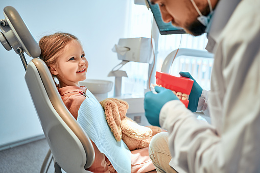 Children's dentistry. A little girl sits in a dental chair and looks at the doctor who shows her a model of the jaw and shows how to brush her teeth.