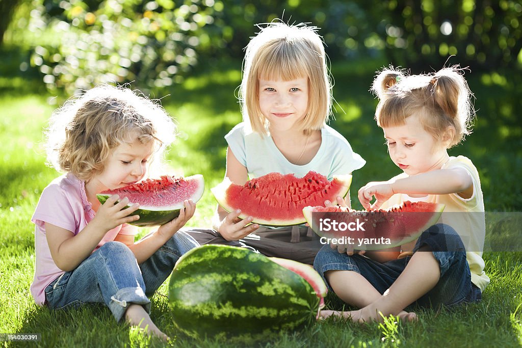 Three blonde girls enjoying a watermelon at a picnic Group of happy children eating watermelon outdoors in spring park Child Stock Photo