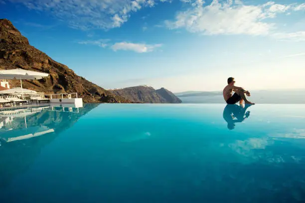 Man sits on edge of infinity pool looking out over view of Santorini caldera, Greece
