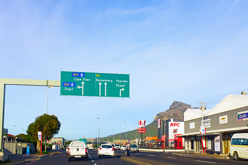 Cape Town, South Africa - July 5, 2023: An overhead direction sign points the way to Paarl, Cape Town, Muizenberg and Paarden Eiland as traffic heads through the suburb of Brooklyn on Koeberg Road in the morning. To the right, Devil's Peak, part of the Table Mountain range, rises above a KFC branch. Pedestrians wait on the center island to cross the road.