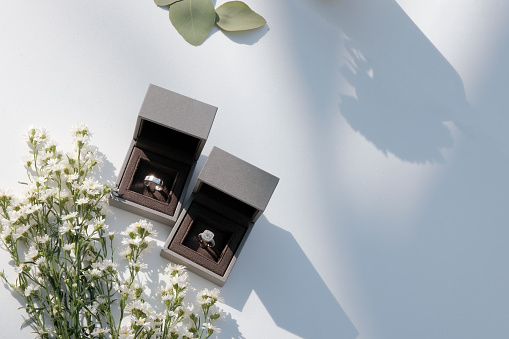 The wedding rings are in boxes on a corner of a white table, decorated with dried flowers, basking in the morning sunlight and shadow in
a blank space.