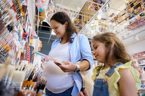 Pregnant woman, mother and her little daughter shopping for school supplies in a school stationery store, choosing felt tip pens, crayons and painting tools. Back to school concept. People. Education